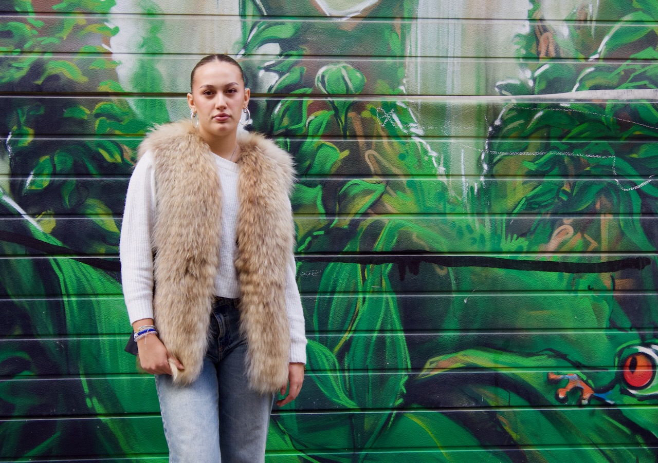 Eine junge Frau steht vor einem Rollbalken. Sie trägt eine blaue Jeanshose und einen weißen Pulli, darüber trägt sie ein klassisches Pelzgilet aus Rotfuchs.